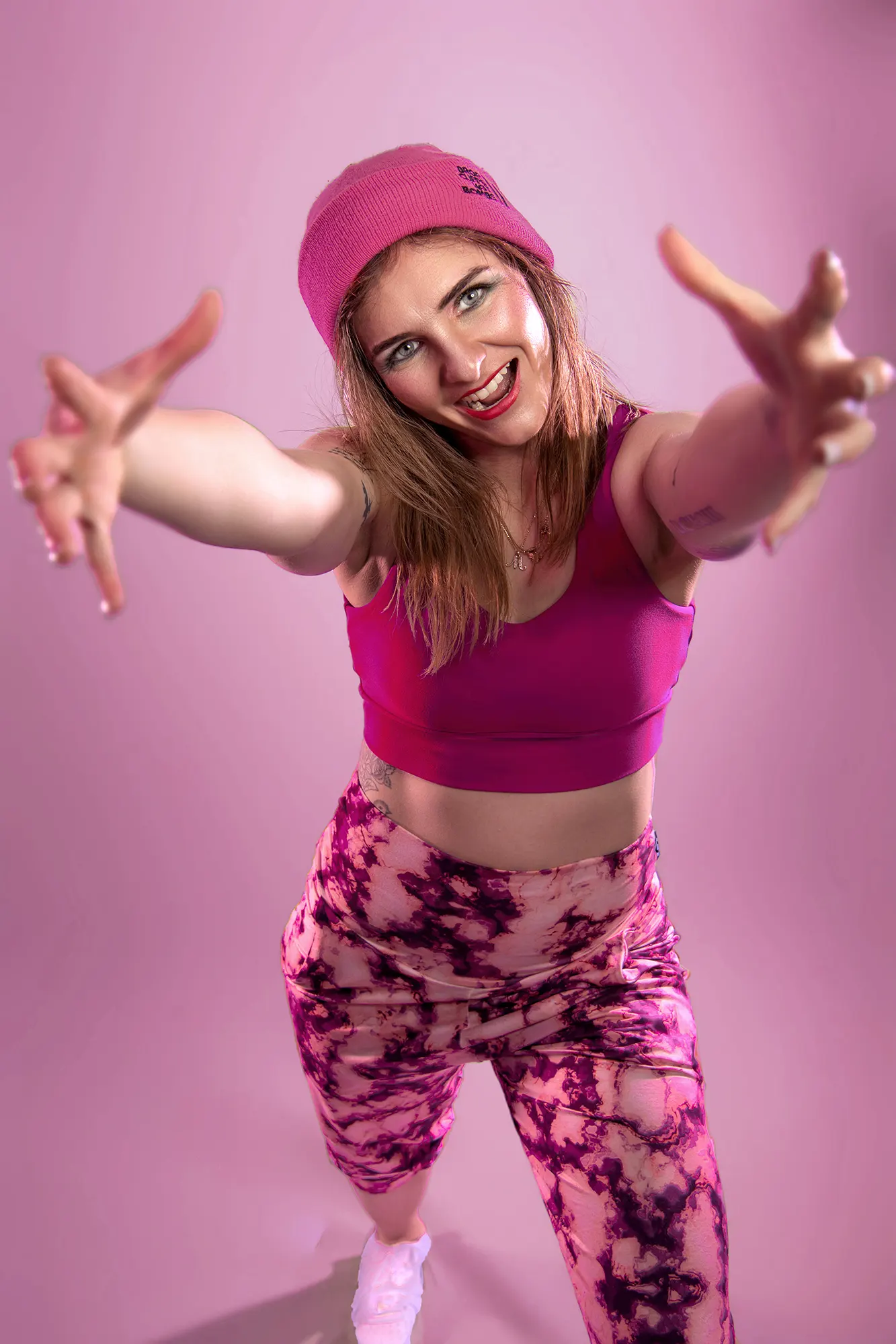 Smiling woman in pink beanie, sports bra, and patterned leggings reaching out, on a pink background
