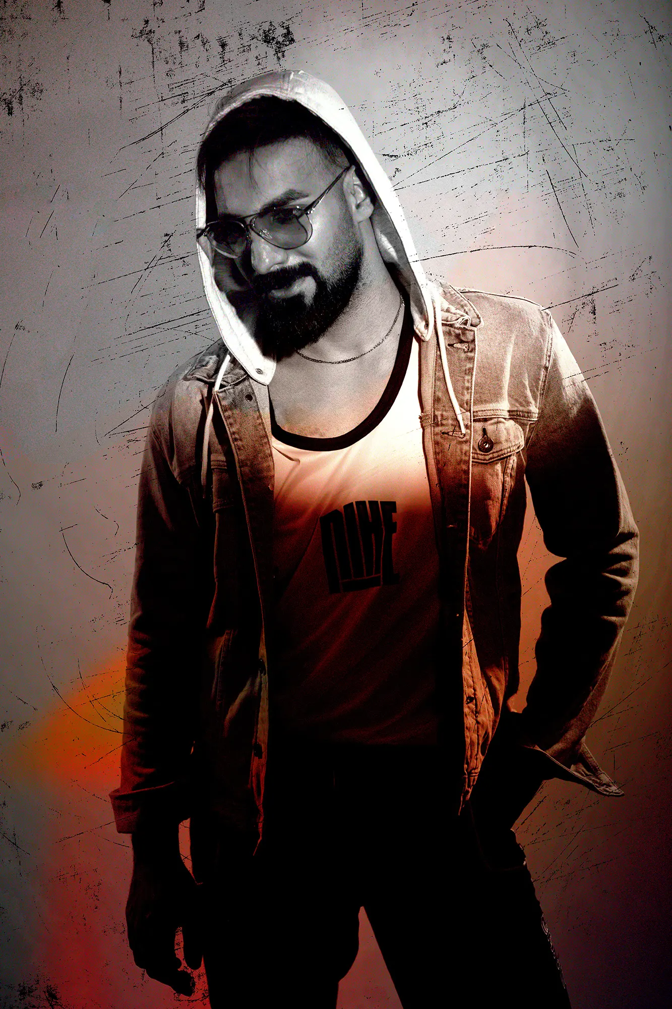 Stylish man with beard wearing hooded denim jacket, aviator sunglasses, and a tank top, against a grunge background.