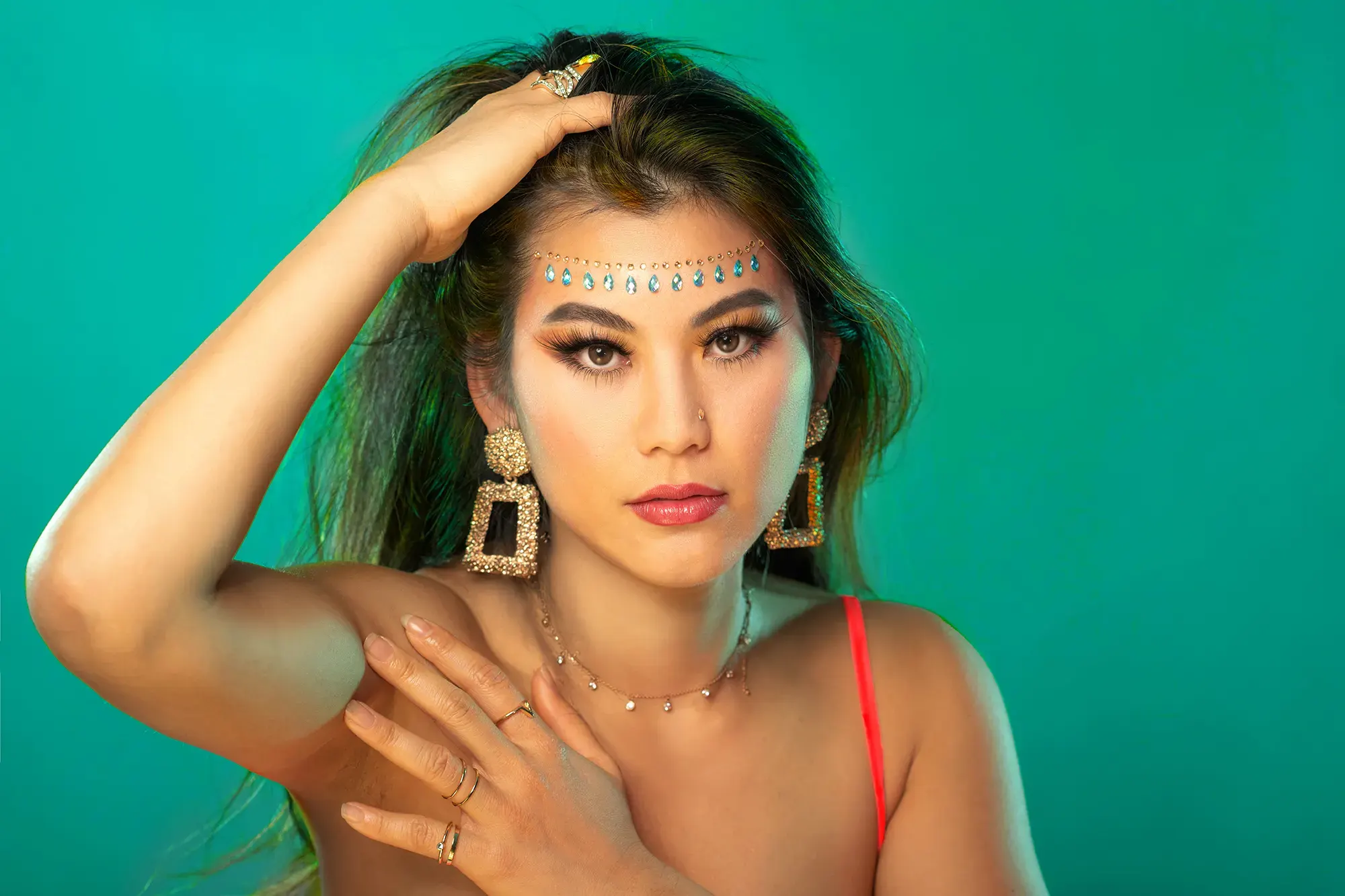 Beautiful woman posing with dazzling jewelry, forehead gems, dramatic makeup, and vibrant teal background