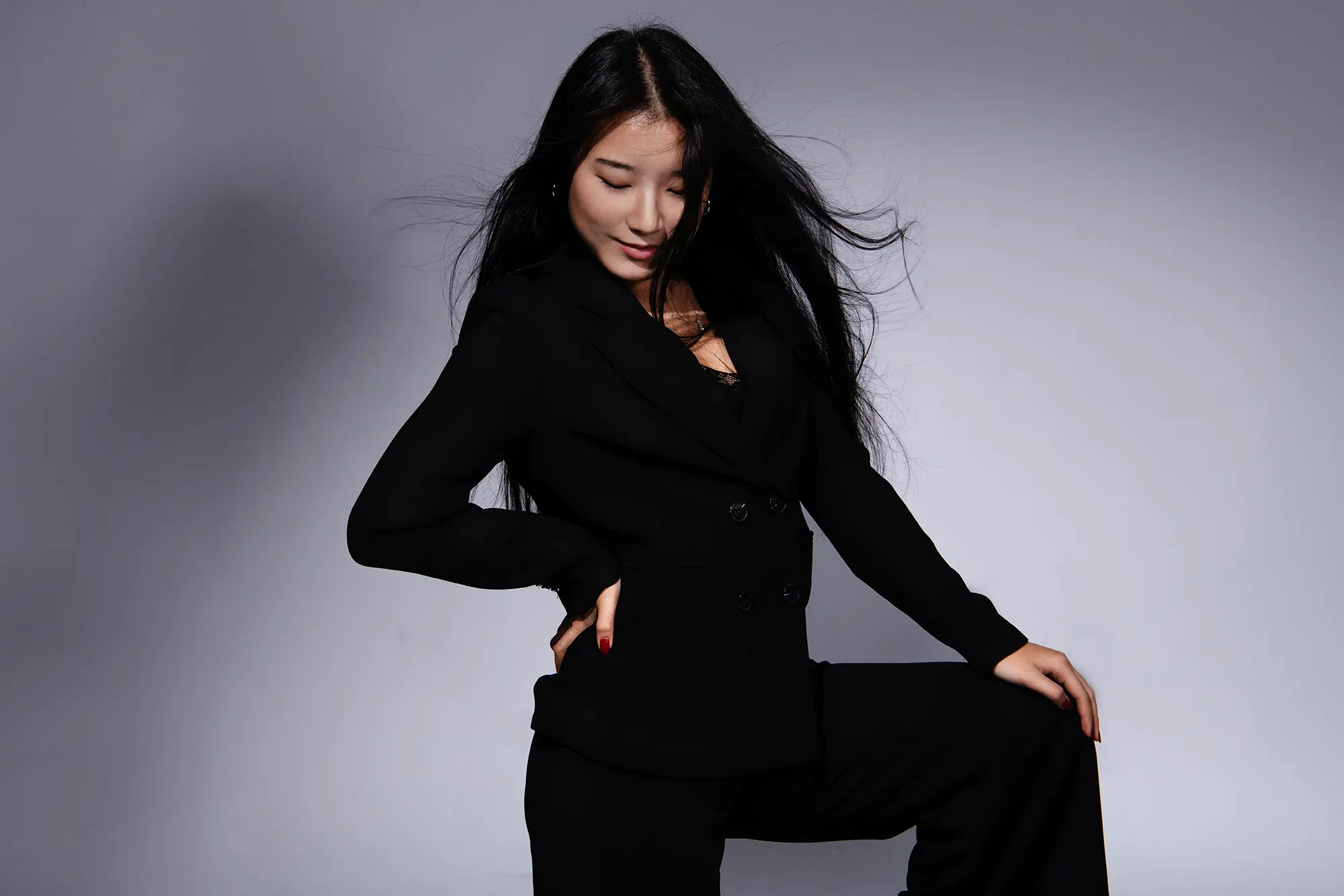 Beautiful Asian woman in a stylish black pantsuit with long flowing hair, posing elegantly against a gray background.