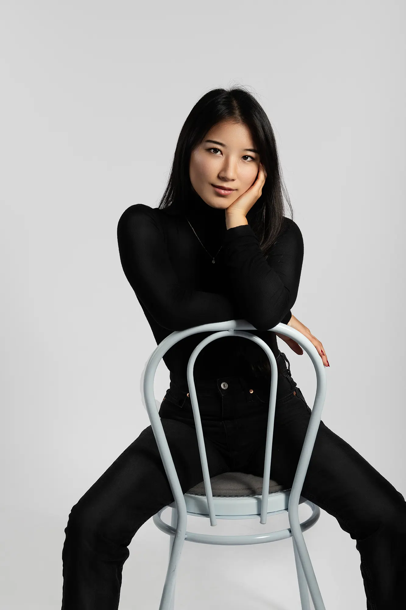 Young Asian woman in black turtleneck and jeans poses on a light blue chair, smiling confidently.